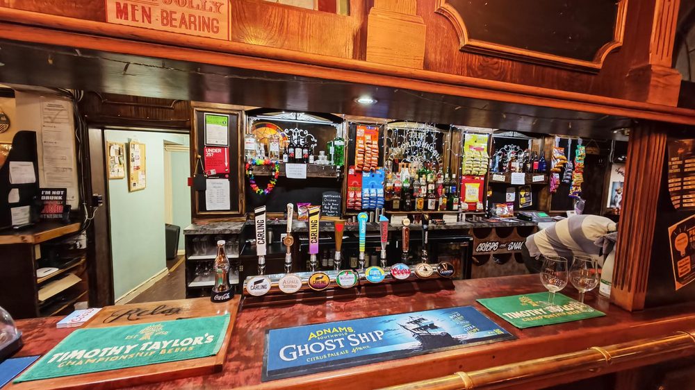 Wide view of the bar showing draught taps, spirits behind the bar, and Timothy Taylor's and Adnams Ghost Ship beer mats