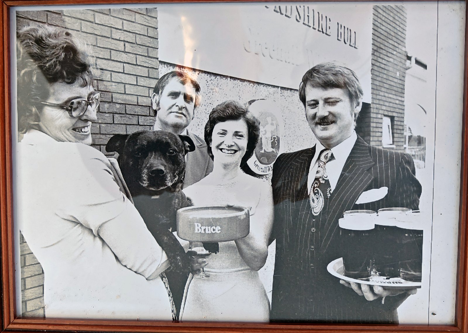 Black and white photo from the opening day, June 1978. Left to right: Shirley Simpson holding Bruce the Staffordshire Bull Terrier and the presentation trophy with 'Bruce' on it, Morris Simpson behind, Noreen Battam, and Graham Battam holding a tray of pints, all standing outside the new pub in front of the Greenall Whitley signage